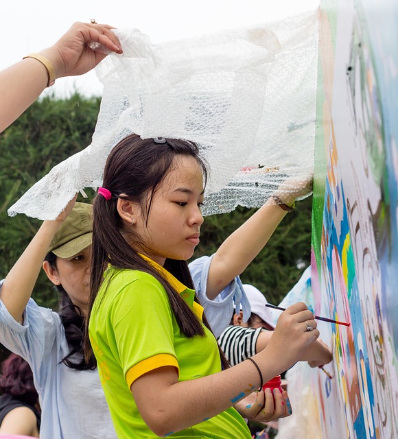 A young girl in a green shirt paints a colorful mural, while an adult holds a protective cover above her to shield from splatters.