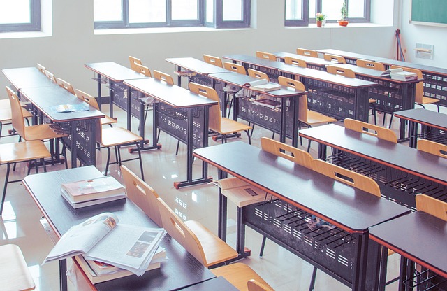 A bright and spacious classroom featuring rows of wooden desks and chairs, with books and materials on some surfaces, and large windows letting in natural light.